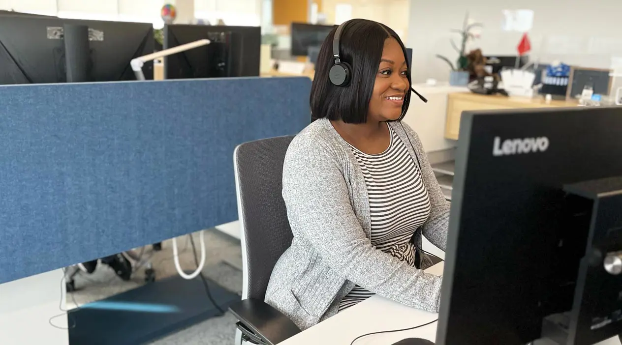 A smiling woman at a computer wearing a headset.